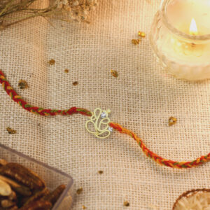 A beautifully handcrafted silver Ganesha Rakhi on a colorful braided thread, placed on a textured beige fabric background with a lit candle and dry fruits nearby.